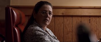 Movie still from “Cop Car” (2015), directed by Jon Watts – An older woman wearing glasses sitting in front of a wooden wall; Close Up shot, Over the shoulder angle