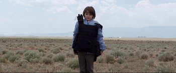 Movie still from “Cop Car” (2015), directed by Jon Watts – A young boy in a field wearing a body armor; Medium shot, Over the shoulder angle