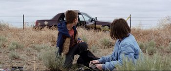 Movie still from “Cop Car” (2015), directed by Jon Watts – Two young boys sitting in the grass near a car; Wide shot, Over the shoulder angle