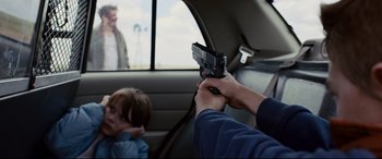 Movie still from “Cop Car” (2015), directed by Jon Watts – A person holding a gun in the back of a car; Medium shot, Over the shoulder angle