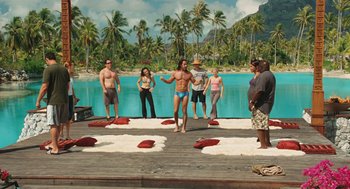 Movie still from “Couples Retreat” (2009), directed by Peter Billingsley – A group of people standing on a dock in front of a body of water and palm trees; Wide shot, High angle