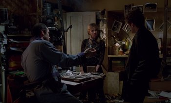 Movie still from “Crash” (1996), directed by David Cronenberg – A group of people standing around a table in a cluttered room; Medium shot, Over the shoulder angle