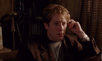 Movie still from “Crash” (1996), directed by David Cronenberg – A man with a brown jacket is sitting down; Close Up shot, Over the shoulder angle
