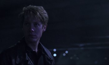 Movie still from “Crash” (1996), directed by David Cronenberg – A man in a black jacket is standing in a dark room; Close Up shot, Low angle