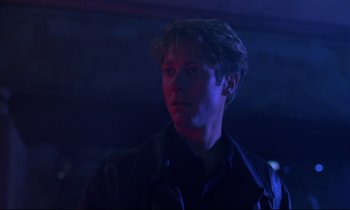 Movie still from “Crash” (1996), directed by David Cronenberg – A man in a black jacket is standing in a dark room; Close Up shot, Low angle