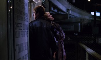 Movie still from “Crash” (1996), directed by David Cronenberg – Two people standing next to each other near a building; Medium shot, Low angle