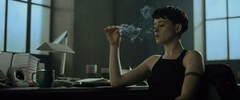 Movie still from “The Girl in the Spider's Web” (2018), directed by Fede Alvarez – A woman sitting at a table with a cigarette in her hand; Medium shot, Low angle