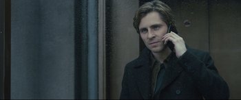 Movie still from “The Girl in the Spider's Web” (2018), directed by Fede Alvarez – A young man holding a cell phone to his ear while standing in front of a window; Close Up shot, Over the shoulder angle