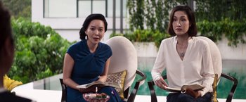 Movie still from “Crazy Rich Asians” (2018), directed by Jon M. Chu – Two women are sitting in a chair and one of them is holding a book; Medium shot, Over the shoulder angle