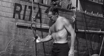 Movie still from “Creature from the Black Lagoon” (1954), directed by Jack Arnold – A shirtless man holding a rope while standing in front of a building; Medium shot, Low angle