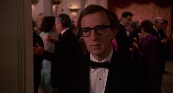 Movie still from “Crimes and Misdemeanors” (1989), directed by Woody Allen – A man wearing a tuxedo and a bow tie in front of a group of people; Close Up shot, Over the shoulder angle