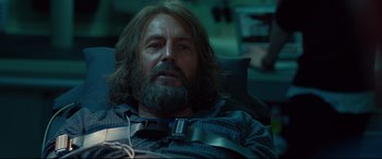 Movie still from “Criminal” (2016), directed by Ariel Vromen – A man with long hair and beard sitting in a chair; Close Up shot, Over the shoulder angle
