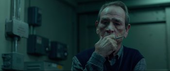 Movie still from “Criminal” (2016), directed by Ariel Vromen – An older man is smoking a cigarette in a dark room; Close Up shot, High angle
