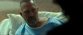 Movie still from “Criminal” (2016), directed by Ariel Vromen – An older man wearing a blue shirt is sitting in a hospital bed; Close Up shot, Over the shoulder angle