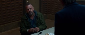 Movie still from “Criminal” (2016), directed by Ariel Vromen – An older man sitting at a table with a microphone; Medium shot, Over the shoulder angle