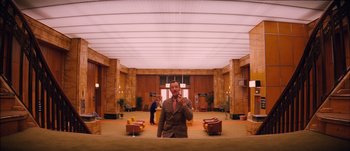 Movie still from “The Grand Budapest Hotel” (2014), directed by Wes Anderson – A man standing in a room with a cell phone to his ear; Wide shot, Low angle
