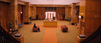 Movie still from “The Grand Budapest Hotel” (2014), directed by Wes Anderson – A woman sitting on a chair in front of a desk in an empty room; Extreme Wide shot, High angle