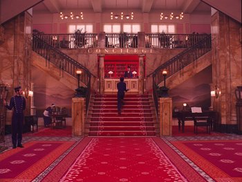 Movie still from “The Grand Budapest Hotel” (2014), directed by Wes Anderson – A man walking up a red staircase in a hotel lobby; Extreme Wide shot, High angle