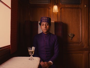 Movie still from “The Grand Budapest Hotel” (2014), directed by Wes Anderson – A man sitting at a table with a glass of wine; Medium shot, High angle