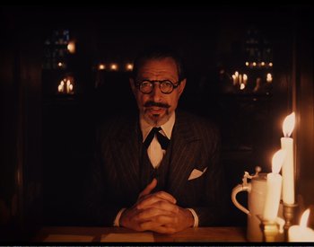 Movie still from “The Grand Budapest Hotel” (2014), directed by Wes Anderson – A man sitting in front of a table with candles; Medium shot, Over the shoulder angle