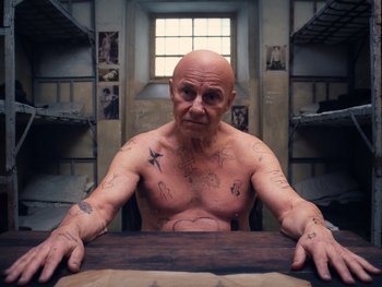 Movie still from “The Grand Budapest Hotel” (2014), directed by Wes Anderson – An old man sitting at a table with tattoos on his arms and chest; Medium shot, High angle
