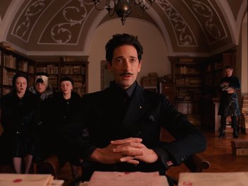 Movie still from “The Grand Budapest Hotel” (2014), directed by Wes Anderson – A man sitting at a table in front of a book case; Medium shot, Low angle