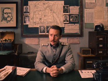 Movie still from “The Grand Budapest Hotel” (2014), directed by Wes Anderson – A man sitting at a table in a room; Medium shot, Low angle