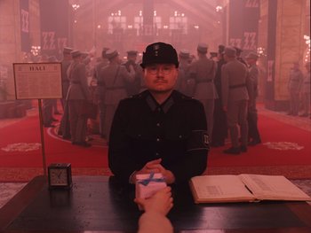 Movie still from “The Grand Budapest Hotel” (2014), directed by Wes Anderson – A man in a uniform sitting in front of a crowd; Medium shot, High angle