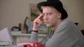 Movie still from “Croupier” (1998), directed by Mike Hodges – A young man wearing a black hat smoking a cigarette in front of a painting; Close Up shot, Low angle