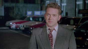 Movie still from “Croupier” (1998), directed by Mike Hodges – A man in a suit and tie standing in front of a car; Close Up shot, Over the shoulder angle