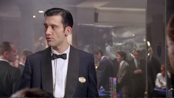 Movie still from “Croupier” (1998), directed by Mike Hodges – A man in a suit and a bow tie in front of a group of people; Medium shot, Over the shoulder angle