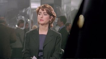 Movie still from “Croupier” (1998), directed by Mike Hodges – A woman sitting in a chair in front of a group of people; Close Up shot, Over the shoulder angle