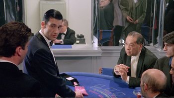 Movie still from “Croupier” (1998), directed by Mike Hodges – A man in a black suit is playing a card game; Medium shot, Over the shoulder angle