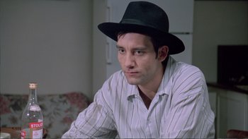 Movie still from “Croupier” (1998), directed by Mike Hodges – A man wearing a black hat sitting in front of a couch; Close Up shot, Over the shoulder angle
