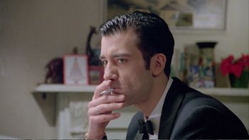 Movie still from “Croupier” (1998), directed by Mike Hodges – A person wearing a suit smoking a cigarette; Close Up shot, Low angle