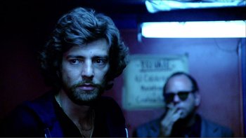 Movie still from “Cruising” (1980), directed by William Friedkin – A man with long hair and a beard standing next to another man; Close Up shot, Low angle