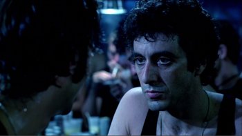 Movie still from “Cruising” (1980), directed by William Friedkin – A person sitting at a table in a room; Close Up shot, Over the shoulder angle