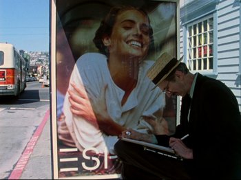 Movie still from “Crumb” (1994), directed by Terry Zwigoff – A man sitting in front of a poster of a smiling woman; Medium shot, High angle