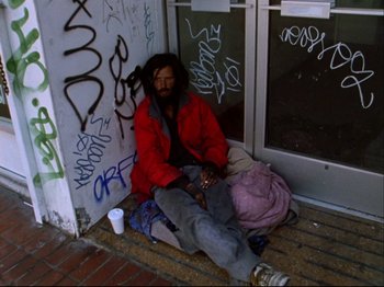 Movie still from “Crumb” (1994), directed by Terry Zwigoff – A man sitting on the ground in front of a building; Wide shot, High angle