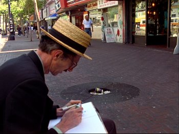 Movie still from “Crumb” (1994), directed by Terry Zwigoff – A man wearing a straw hat writing on a piece of paper on the sidewalk; Wide shot, High angle
