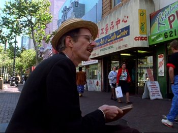 Movie still from “Crumb” (1994), directed by Terry Zwigoff – An older man sitting on the side of the street while using a cell phone; Medium shot, Low angle