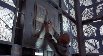 Movie still from “Cube” (1997), directed by Vincenzo Natali – A bald man in a gray jacket is working on a door; Medium shot, Low angle