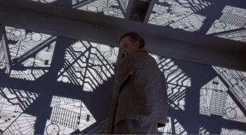 Movie still from “Cube” (1997), directed by Vincenzo Natali – A man standing in front of a building with a shadow on the wall; Medium shot, High angle
