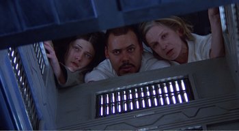 Movie still from “Cube” (1997), directed by Vincenzo Natali – A group of three people looking out of an elevator; Medium shot, Overhead angle