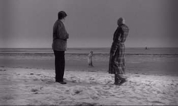 Movie still from “Cul-de-sac” (1966), directed by Roman Polanski – Two people standing on the beach looking at a dog; Extreme Wide shot, High angle