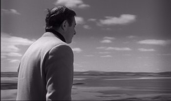 Movie still from “Cul-de-sac” (1966), directed by Roman Polanski – A black and white photo of a man looking out at the ocean; Medium shot, Over the shoulder angle
