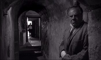 Movie still from “Cul-de-sac” (1966), directed by Roman Polanski – A man in a suit and tie standing in an alley; Medium shot, Low angle