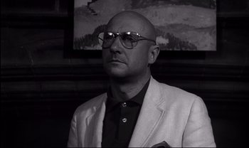 Movie still from “Cul-de-sac” (1966), directed by Roman Polanski – A bald man wearing a white jacket and glasses; Close Up shot, Low angle