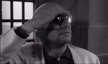 Movie still from “Cul-de-sac” (1966), directed by Roman Polanski – A man in a suit and sunglasses saluting with one hand; Close Up shot, Low angle