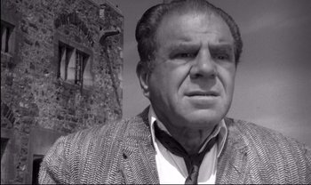 Movie still from “Cul-de-sac” (1966), directed by Roman Polanski – An older man wearing a jacket and a tie in front of a stone building; Close Up shot, Low angle